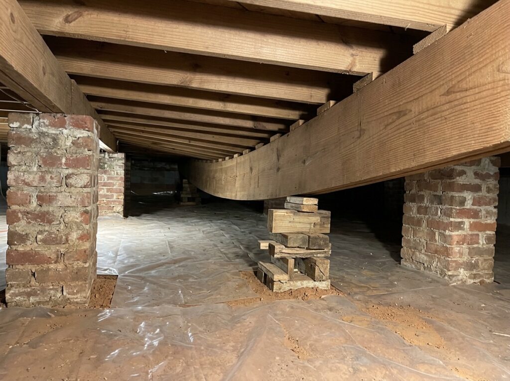 Crawl space girder beam bowing down between widely spaced brick piers, shimmed with wood blocks